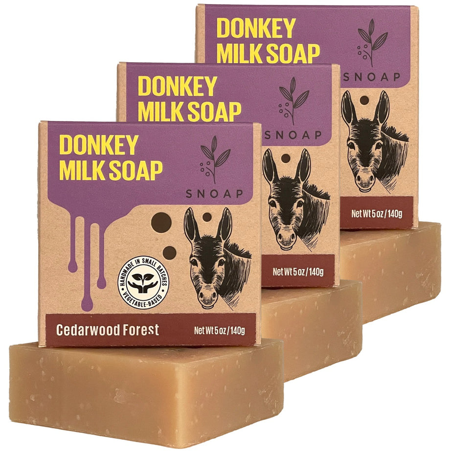 Three bars of cedarwood forest donkey milk soap with a light brown color are stacked in a staggered formation, each below its cardboard box packaging. The boxes are decorated with a sketch of a donkey and the text 'Donkey Milk Soap' and 'SNOAP'.