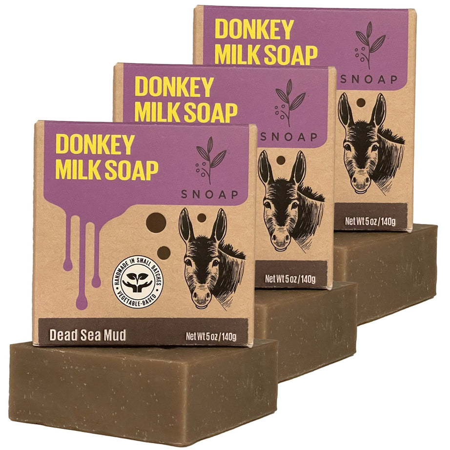 Three bars of dead sea mud donkey milk soap with a dark brown color are stacked in a staggered formation, each below its cardboard box packaging. The boxes are decorated with a sketch of a donkey and the text 'Donkey Milk Soap' and 'SNOAP'.