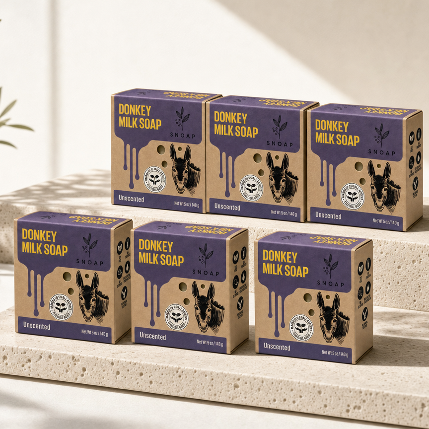 SNOAP 6-Box Half-Year Reserve: Six Pure Donkey Milk Soap unbleached kraft packaging boxes arranged in an architectural stepped two-tier pantry reserve composition on honed ivory travertine stone plinths with soft morning sunlight.